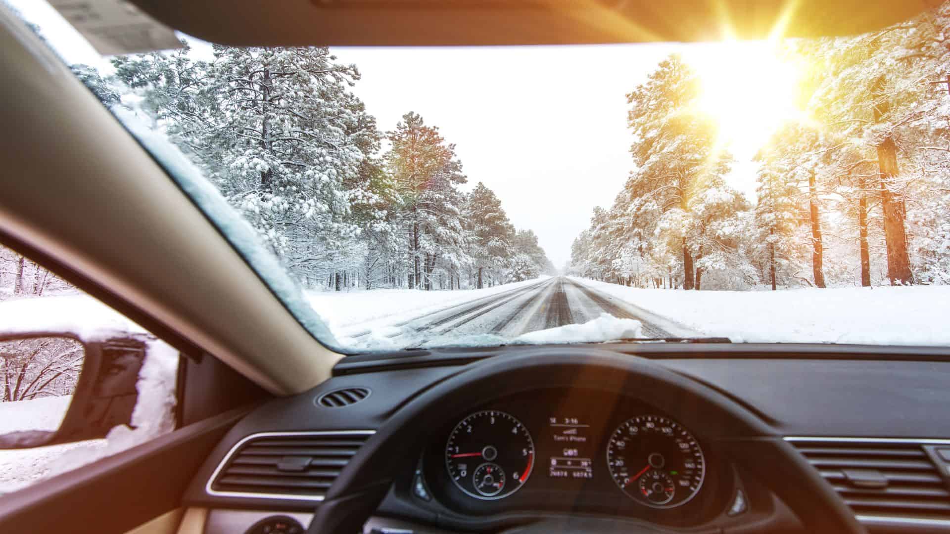 Snowy winter road driving from inside a car with sunlight and snow-covered trees.