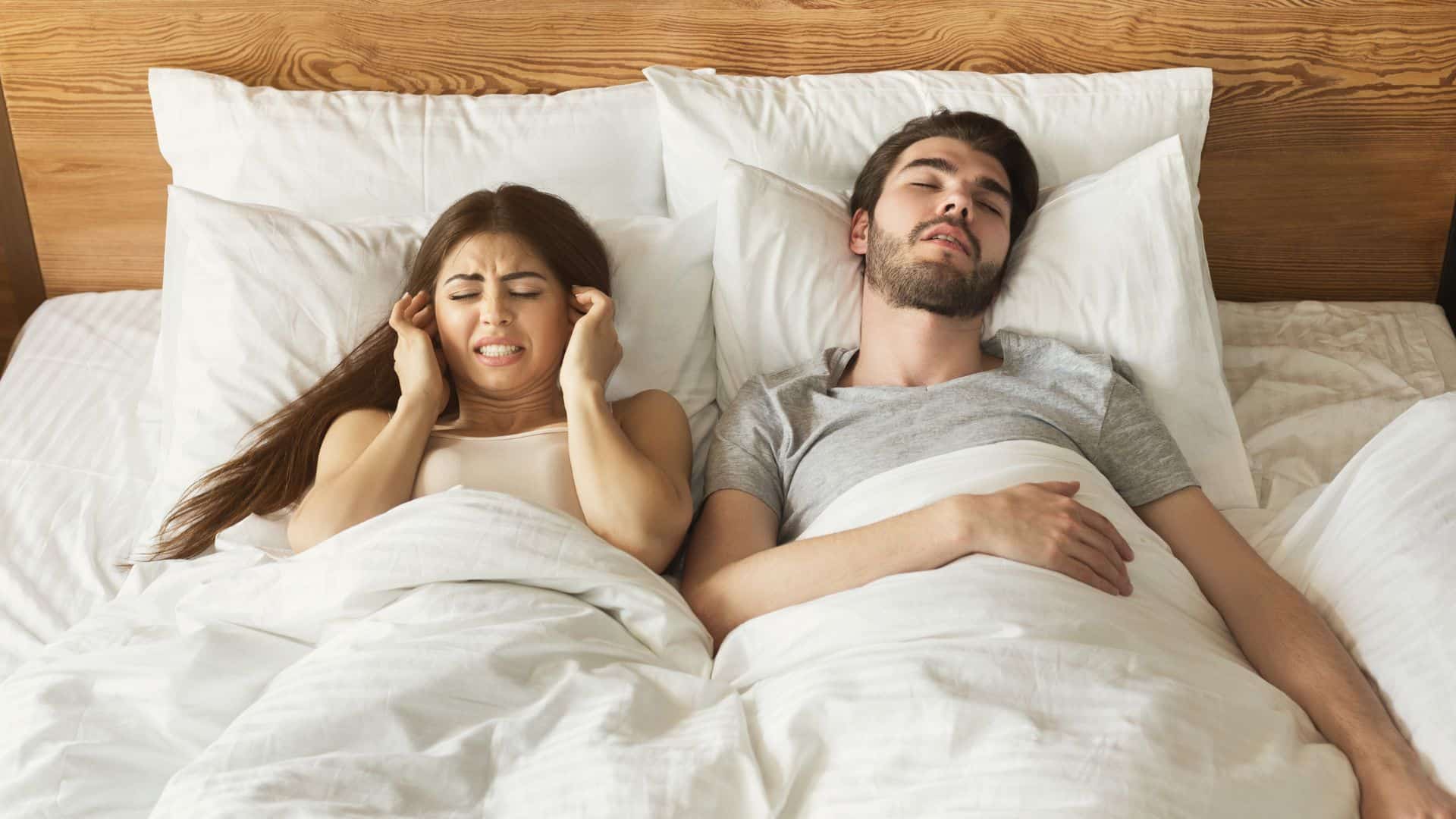 Woman plugging her ears laying in bed next to snoring husband