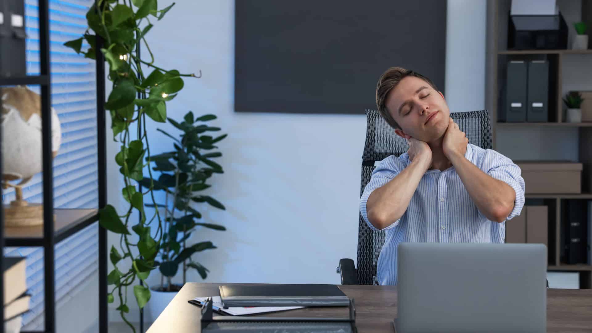 A tired man sitting at his desk with his hands rubbing the sides of his neck