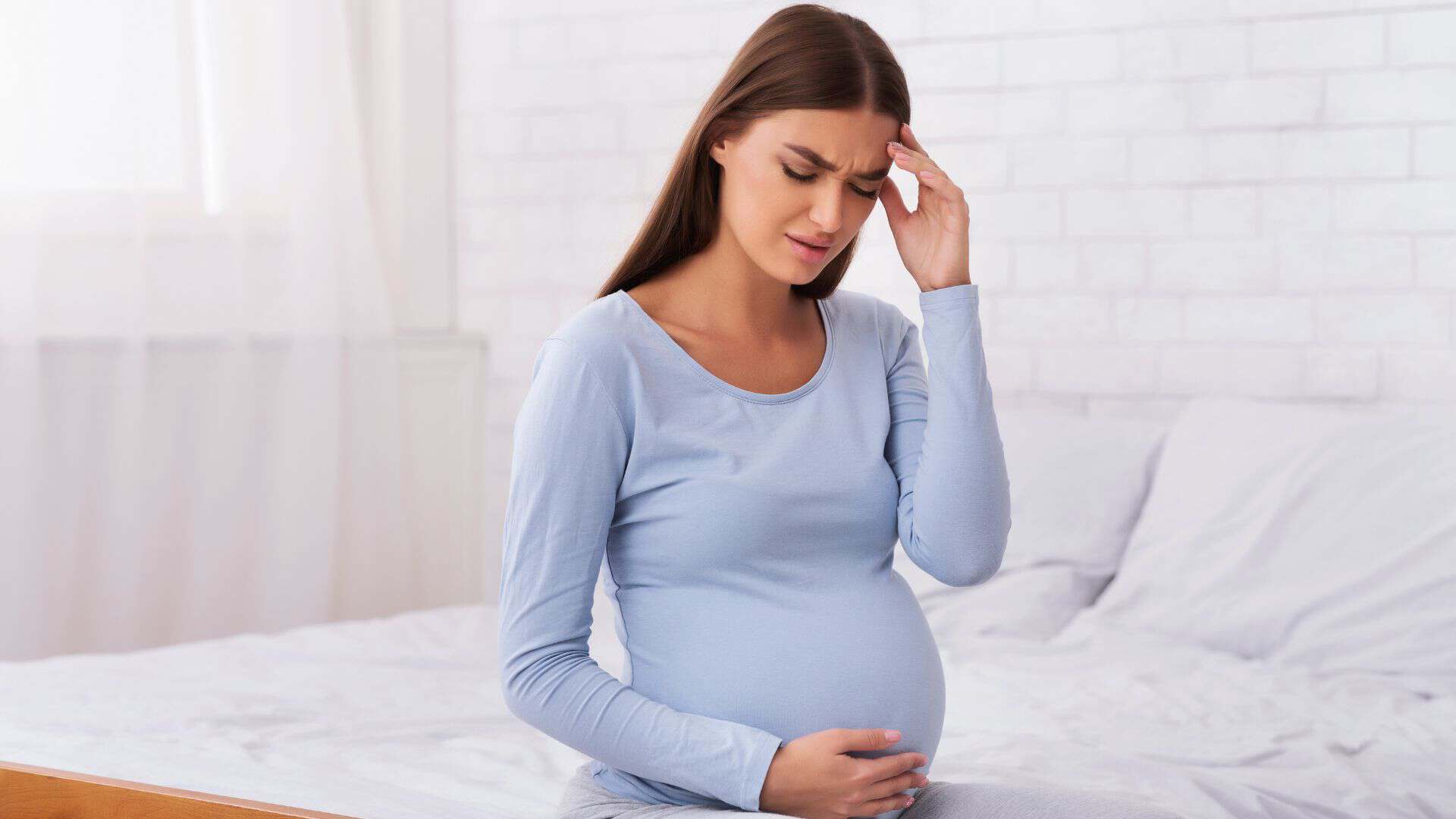 A frustrated pregnant woman sitting on the bed holding her belly.