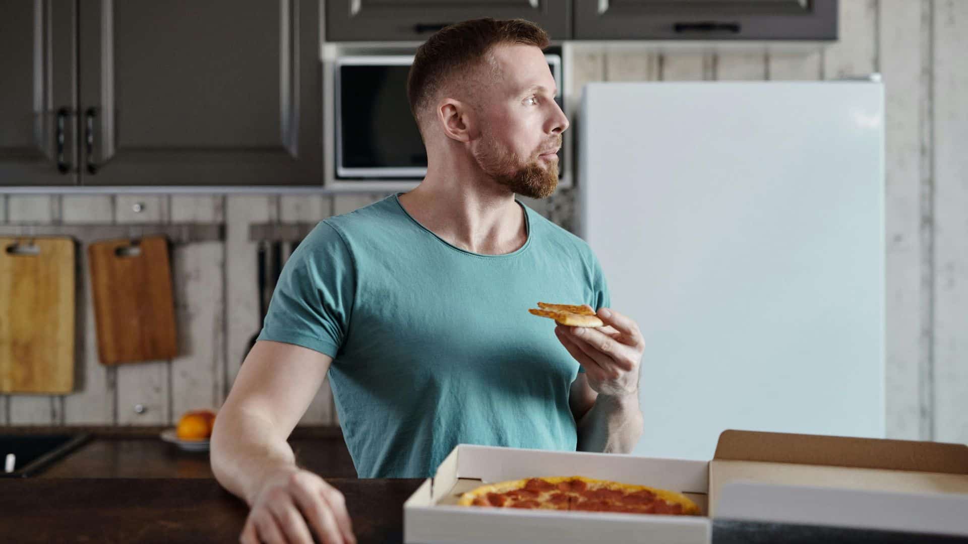 A man standing in his kitchen eating pizza