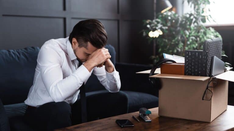 A young man is sitting, stressed, with a box of his belongings on the table.
