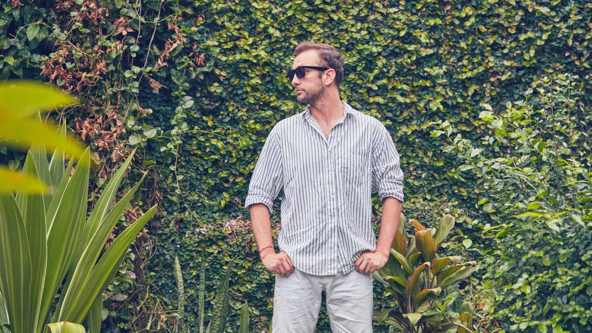 A man wearing sunglasses poses in front of greenery