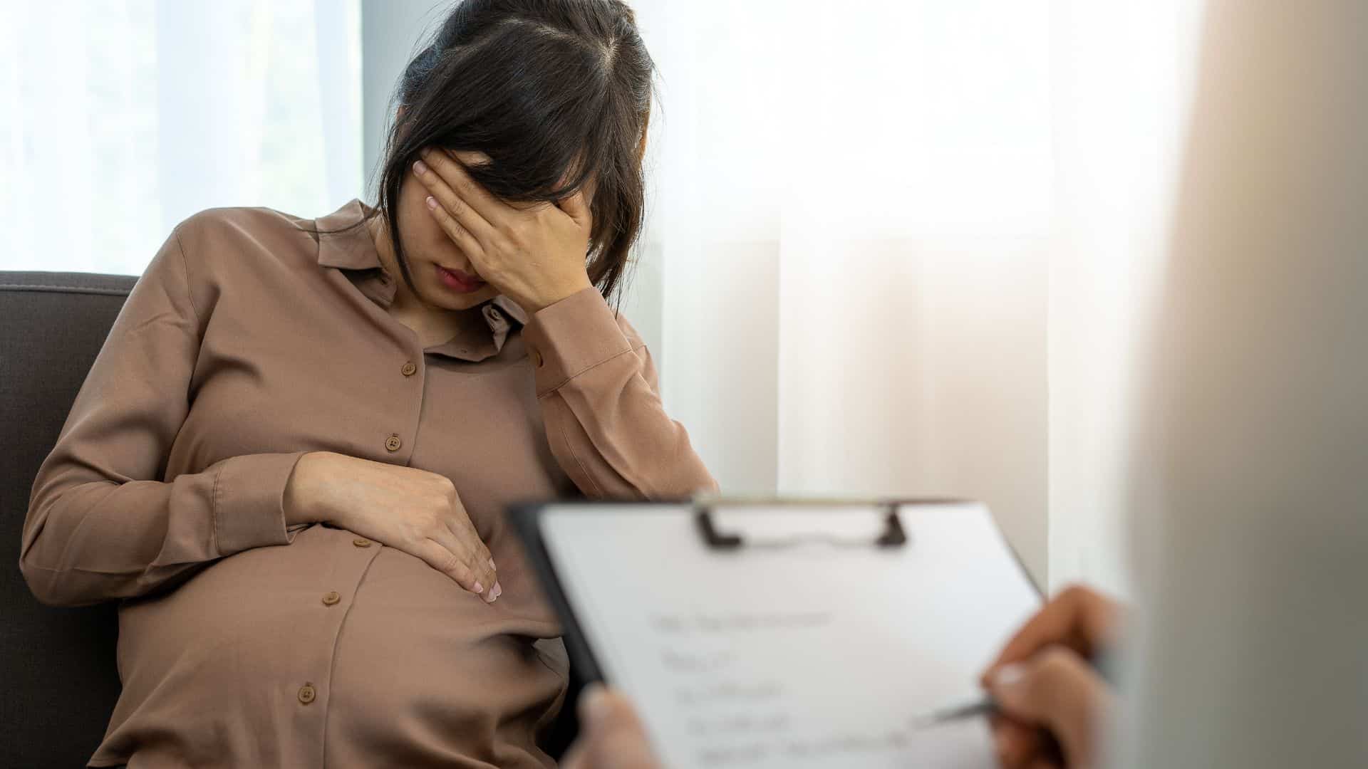 A pregnant woman looking stressed, holding her belly with her hand on her head.